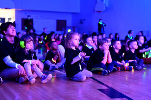Students watching the concert, all a-glow with glowstick necklaces and bracelets in a black lit gym.
