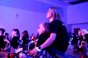 Students and staff watching the concert, all a-glow with glowstick necklaces and bracelets in a black lit gym.